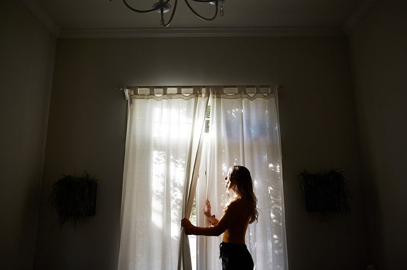 A Christian woman standing in a dark room, peeking through curtains to look outside
