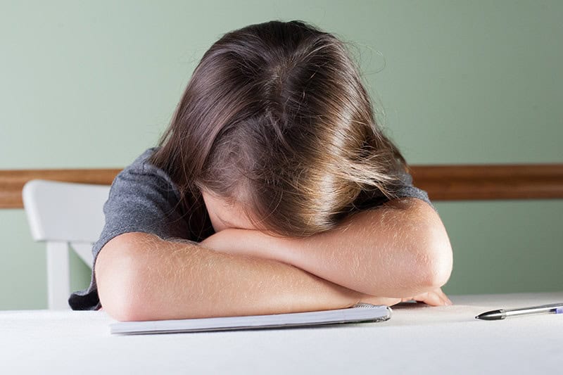 a young girl at a desk with her head down on her folded arms in discouragement