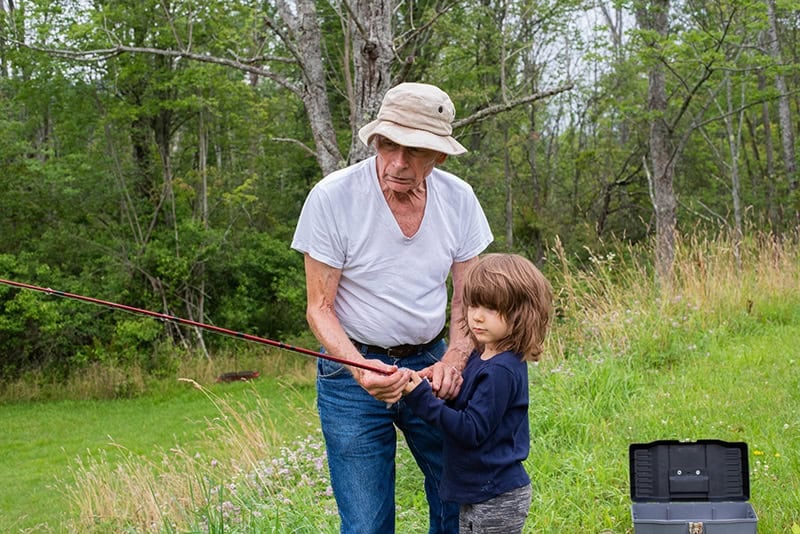 A tackle box sits open on the ground beside a Christian grandfather wearing a fishing hat and teaching his grandson how to fish by helping him hold a red fishing pole as a way to pass down your faith to the next generation