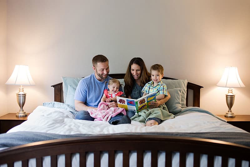 A Christian family&mdash;dad, mom, toddler boy and toddler girl&mdash;snuggling in the parents' bed as they enjoy a colorful children's book together