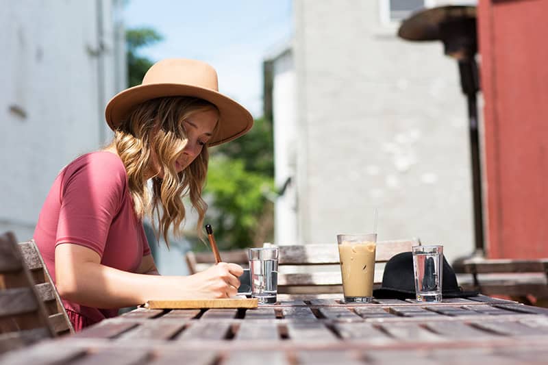 A Christian woman sitting at a table outdoors wearing a sun hat and journaling God's faithfulness