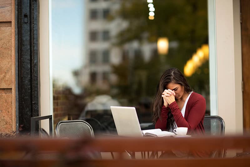 A Christian woman praying with her Bible and laptop open at an outdoor cafe