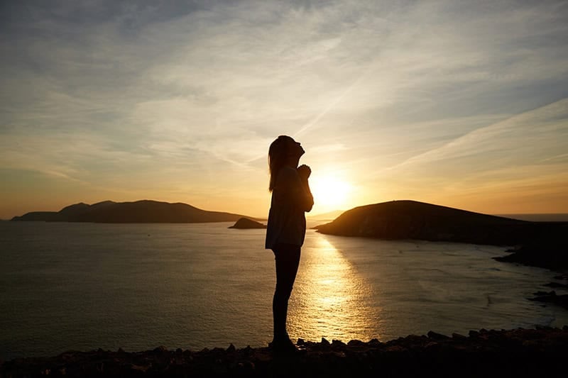 A Christian woman standing on the shore at sunset with her head lifted in prayer to transform anxiety into peace