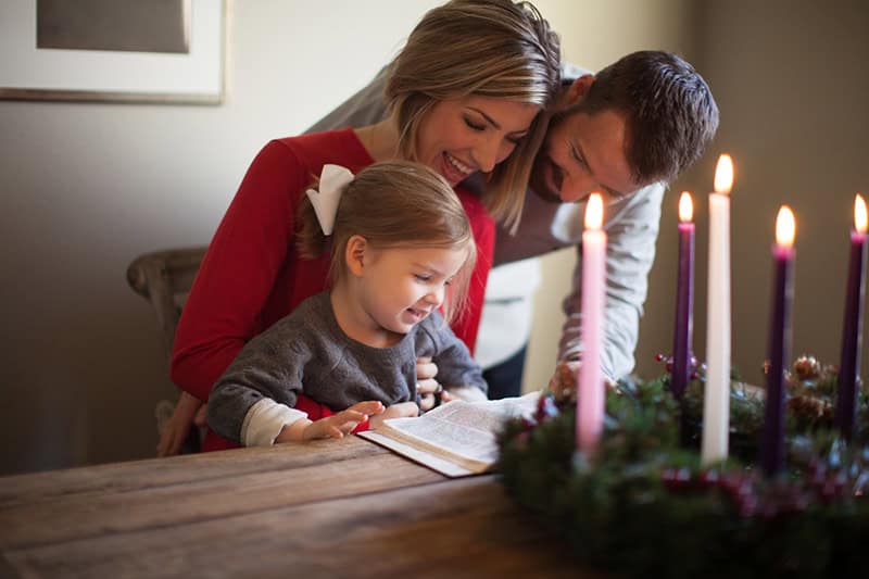 A Christian family gathered around the table with an open Bible and an Advent wreath and five lit candles to talk about the meaning of Advent