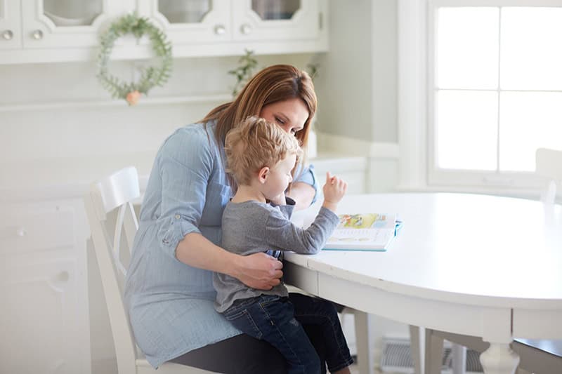 A Christian mom holding her toddler in her lap at the dining table, reading a children's book to him