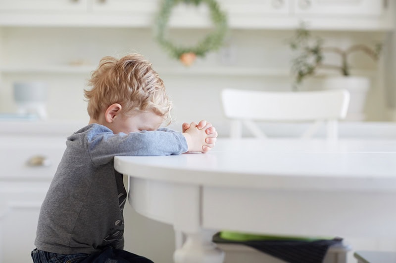 A toddler sitting at a white dining table with his head down and his hands folded as he learns a lesson about obedience and God's mercy