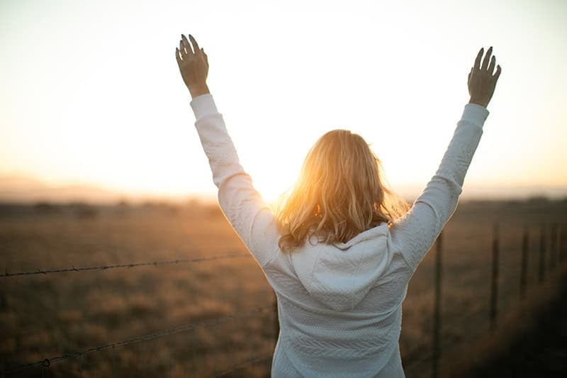 A Christian woman facing the sunset with her arms raised in surrender