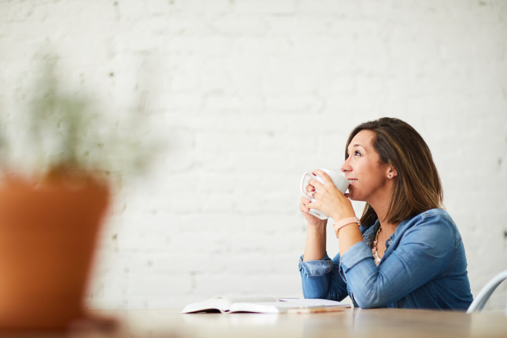 A Christian woman smiling as she meditates on the Bible at the table with a mug in her hands