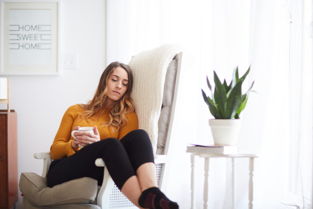 A Christian woman enjoying the rest that comes from rhythms of grace as she sits in a rocking chair with a mug in her hand and her eyes closed