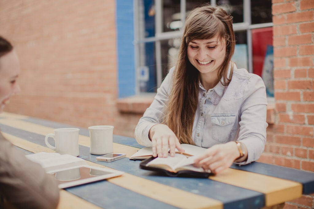 A Christian woman grinning with delight as she studies the Bible with a friend outdoors at a blue and yellow picnic table