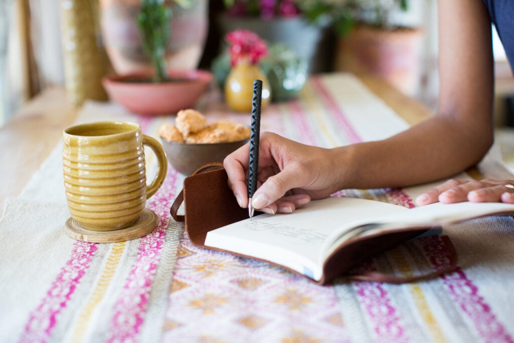 A Christian woman writing in a gratitude journal with a mug and a snack beside her at table full of colorful succulent plants
