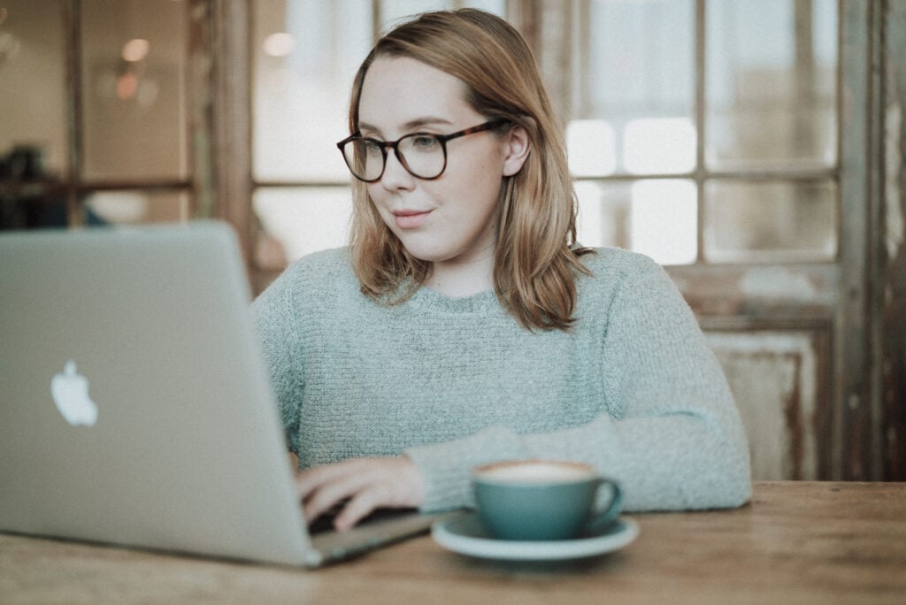 A Christian young woman wearing glasses typing on a laptop with a mug beside her on a rustic table
