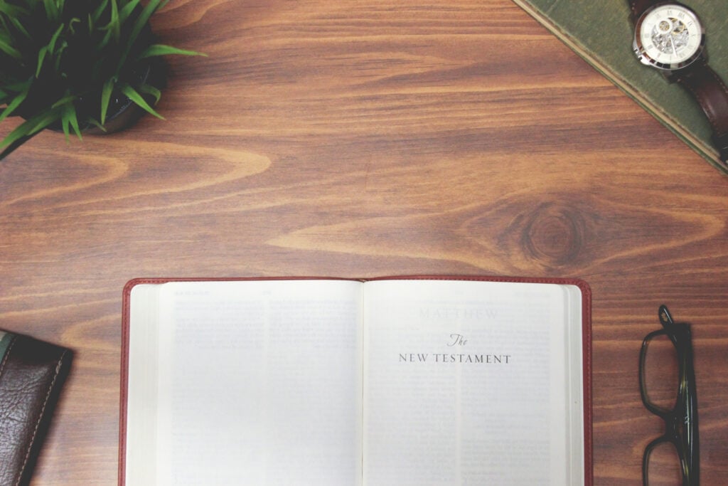 A Bible open to the New Testament on a wooden desk with a pair of glasses, a watch, and a leather notebook beside it