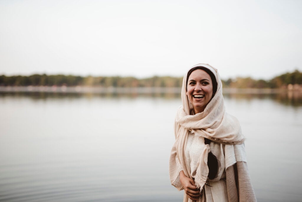 A woman dressed in ancient biblical clothing standing near a calm lake with joy on her face