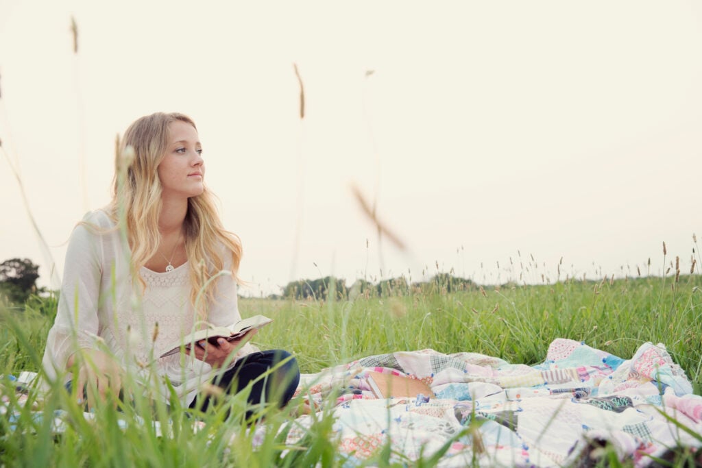 A Christian woman contemplating the power of praise as she holds her open Bible while sitting on a patchwork picnic blanket in a green meadow