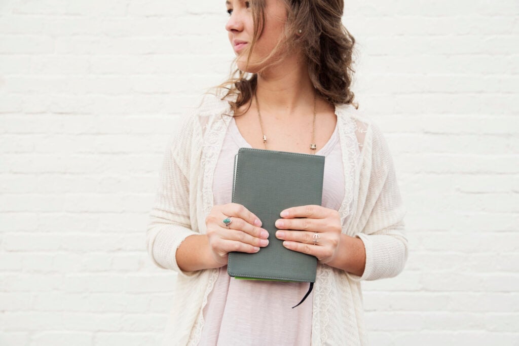 A Christian young woman against a white brick backdrop, holding a Bible in both hands while waiting on God