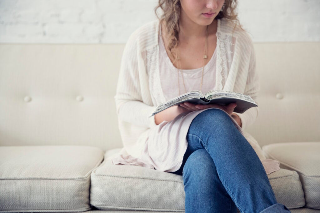 A young Christian woman sitting on an off-white couch, reading Bible verses for waiting on God