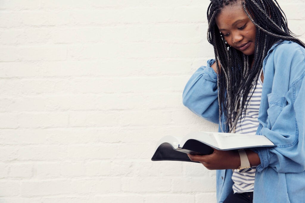 A young Christian woman standing in front of a white brick wall, reading Bible verses for waiting on God