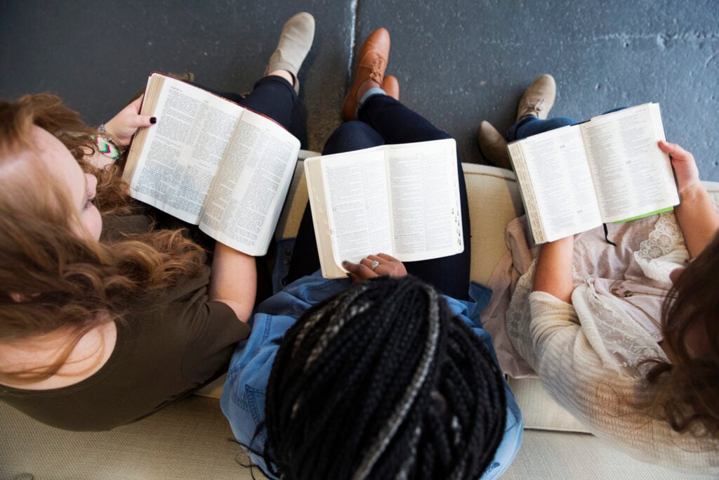 An overhead shot of three young Christian woman sitting side-by-side on a couch, reading the Bible together