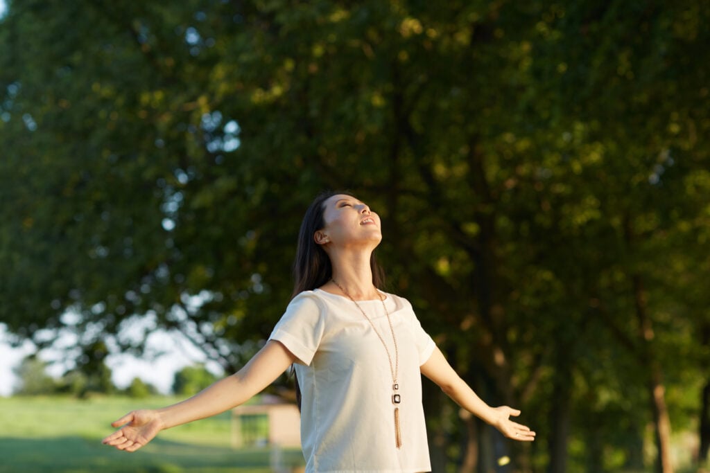 A Christian woman outdoors with her head tilted toward the sun and her arms open in surrender to God