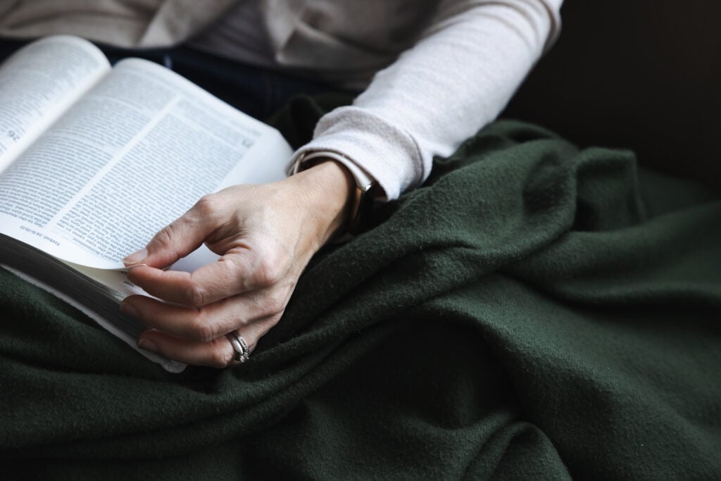 A Christian woman snuggled under a cozy dark green blanket with her Bible open in her lap