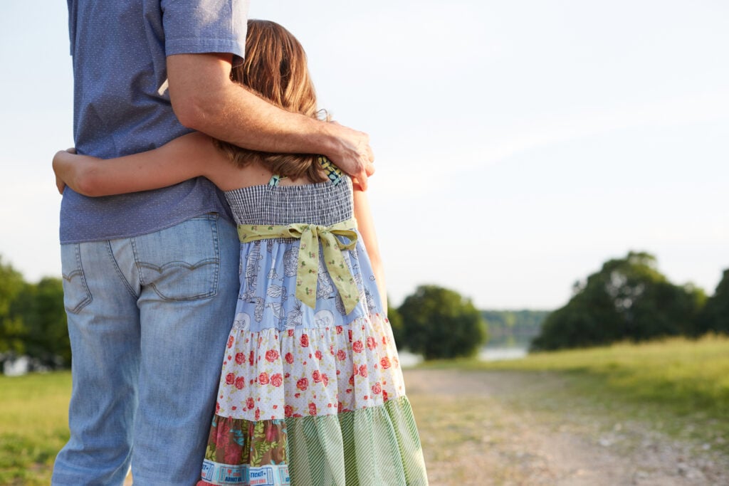 A Christian father with his arm wrapped around his young daughter as they look across a dirt road toward a lake
