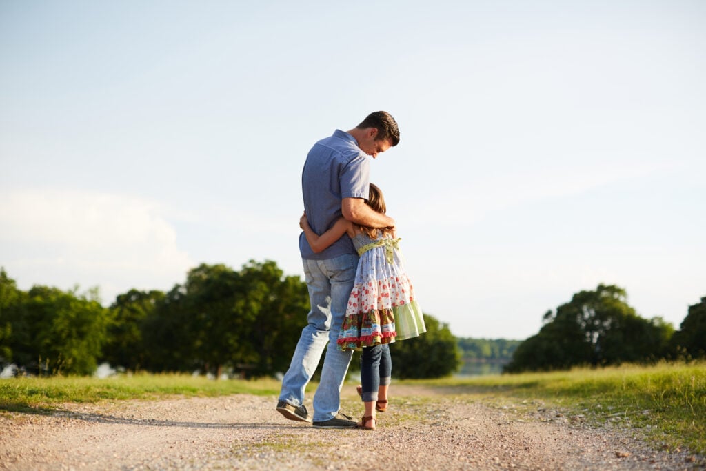 A Christian father hugging his young daughter as they walk along a dirt road, illustrating God's kindness in the prodigal son parable
