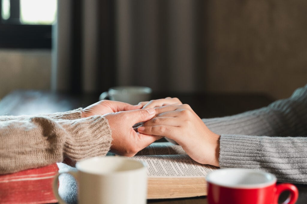 Two Christian women holding hands in prayer across a table with an open Bible and coffee mugs after reading Philippians 4:4&ndash;9 to calm anxiety