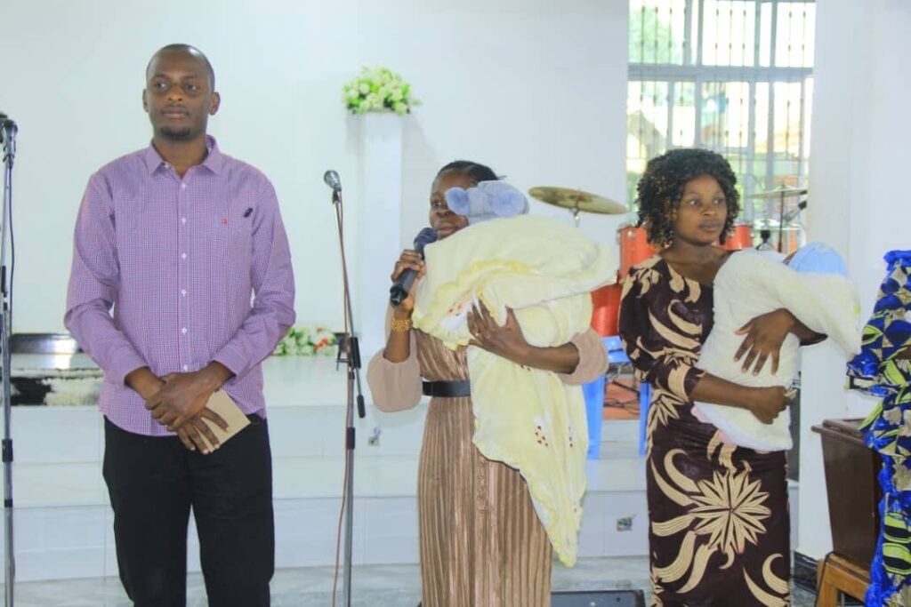 A Christian Bible teacher and church deacon in Tanzania standing in front of the church alongside two women holding babies