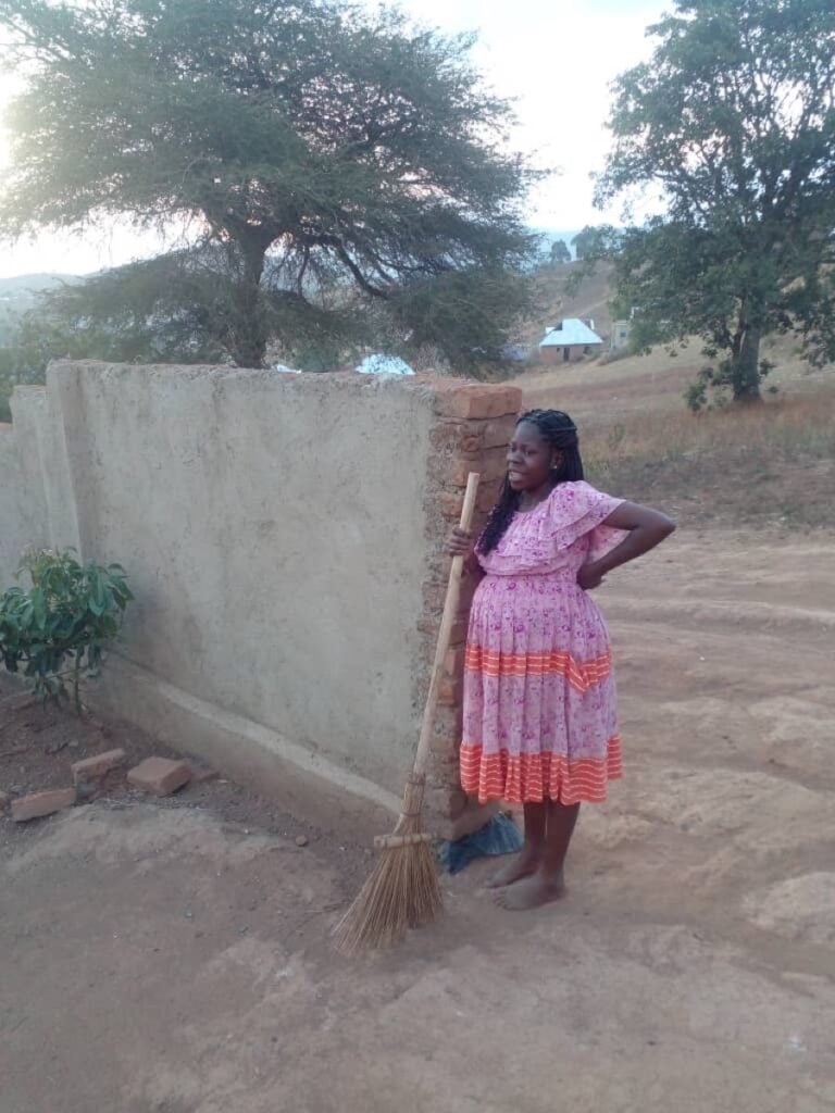 A pregnant Christian woman in Tanzania, standing in her dirt yard with a straw broom, waiting on God in difficult times
