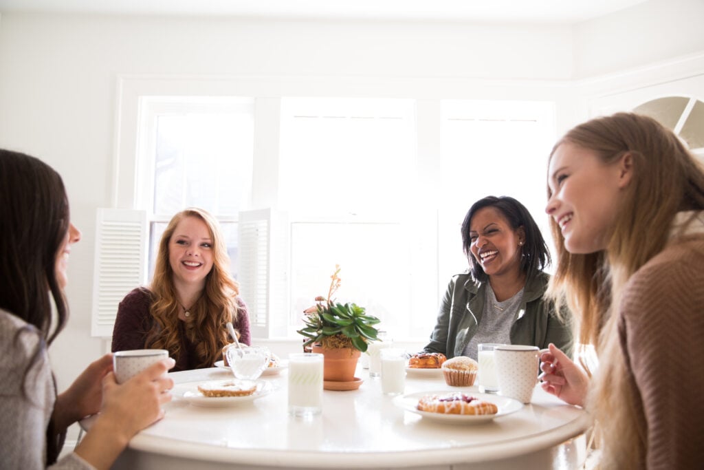 A group of four smiling Christian woman gathered around a table, drinking glasses of milk, mugs of coffee, and enjoying great conversation and delicious pastries