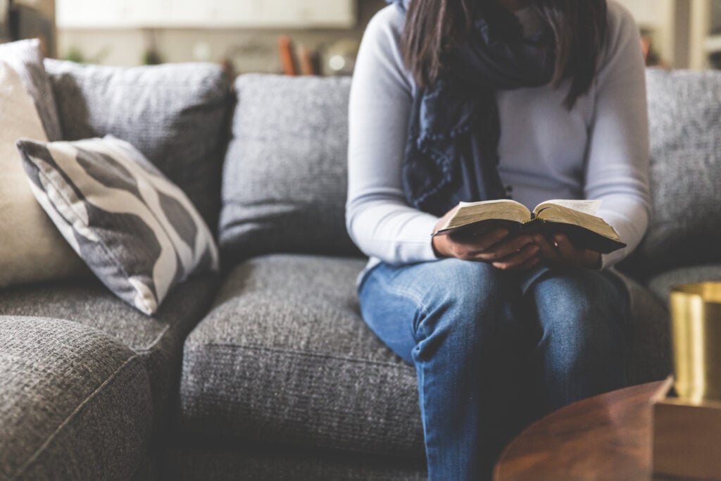 A Christian woman sitting on a grey couch, holding an open Bible as she learns how to stop worrying and trust God