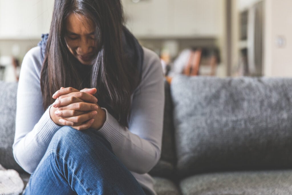 A Christian woman sitting on a grey couch with her legs crossed and her hands folded on her lap in prayer as she practices how to stop worrying and trust God