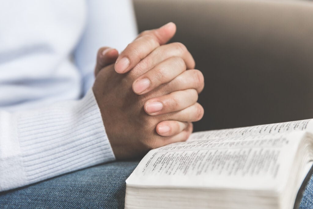 A Christian woman with her hands folded on top of the open Bible on her lap