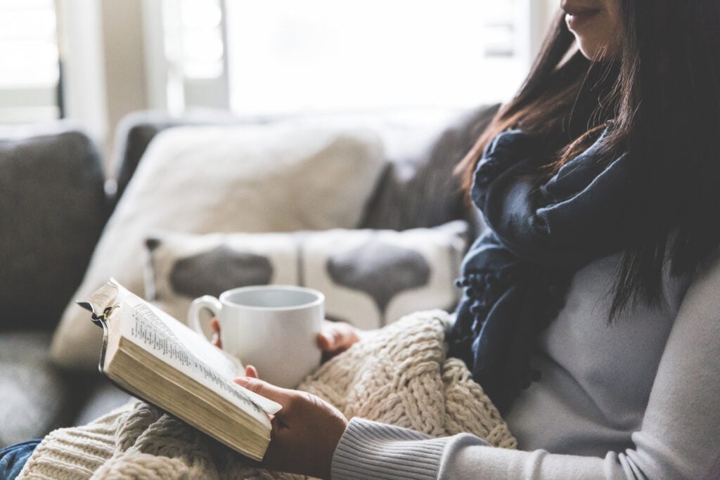 A Christian woman on a grey couch with a blanket on her lap, holding a mug and an open Bible