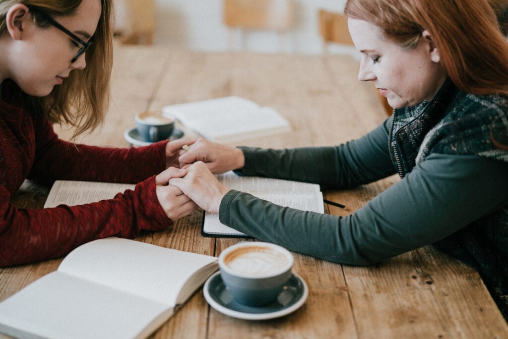Two Christian women holding hands in prayer across a table with open Bibles
