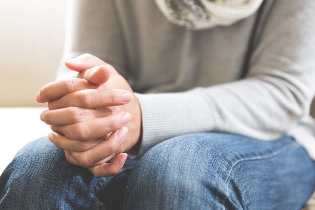 A Christian woman wearing jeans, a grey sweater, and a white scarf, sitting on a white couch with her hands folded in her lap