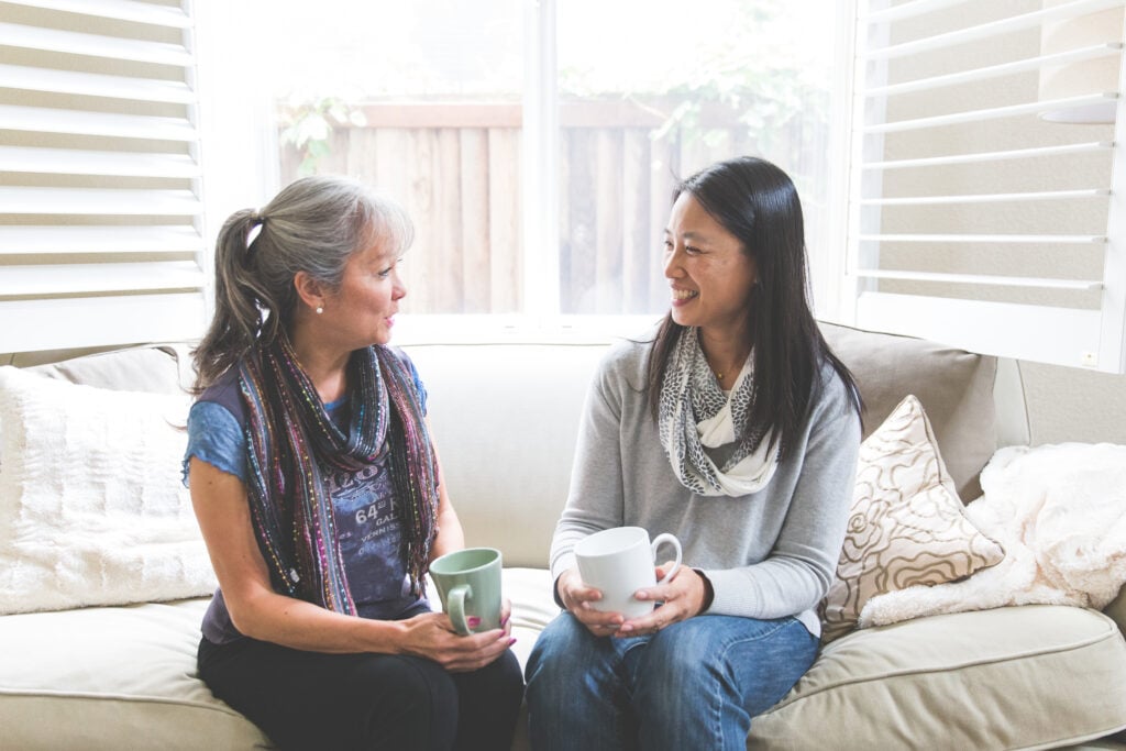 Two Christian woman sitting beside each other on a white couch, holding coffee mugs, and smiling as they converse about the live at peace with everyone meaning