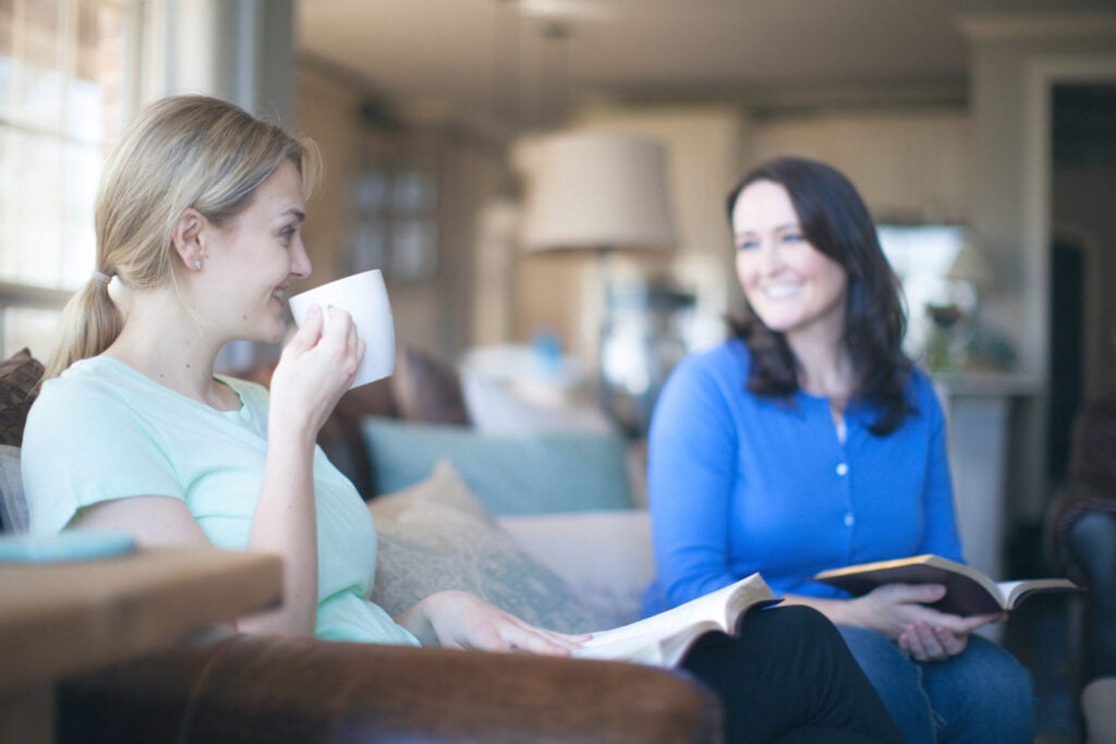 Two Christian women sitting on a couch with open Bibles to learn the difference between peacemaker vs peackeeper, as one woman sips from a coffee mug