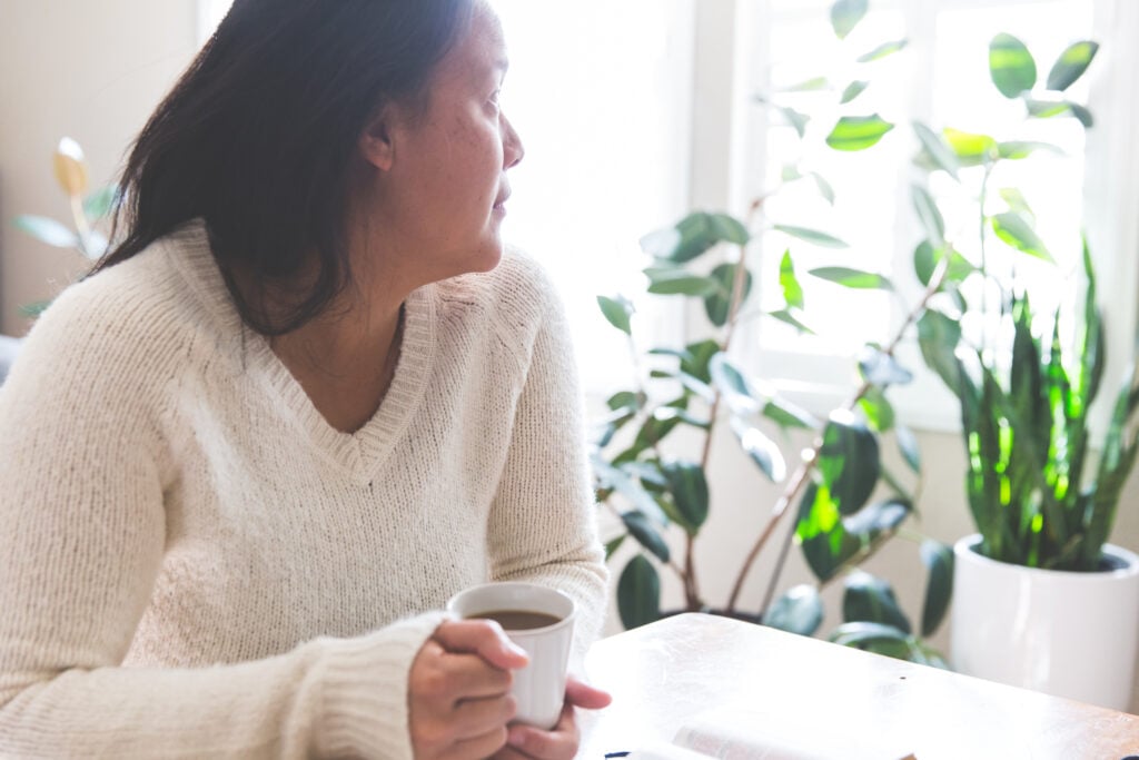 A Christian woman sitting at a table with a mug of coffee, looking out her window while trusting God when He is silent