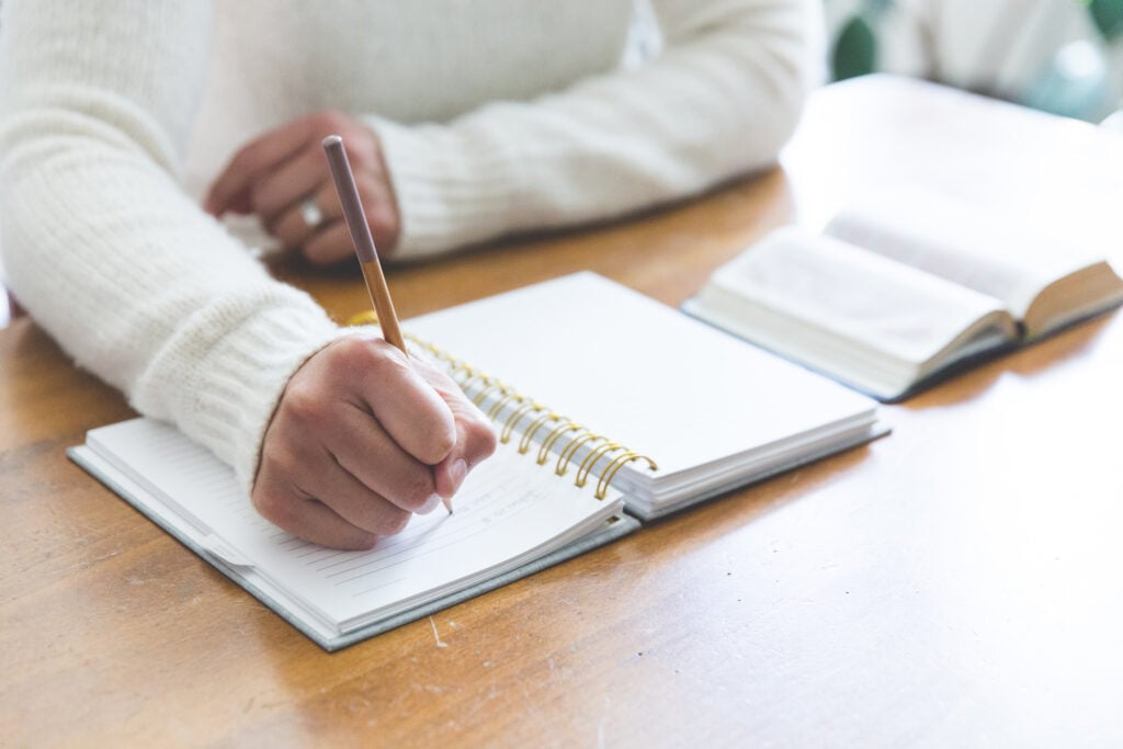 A Christian woman journaling in a notebook with an open Bible beside it