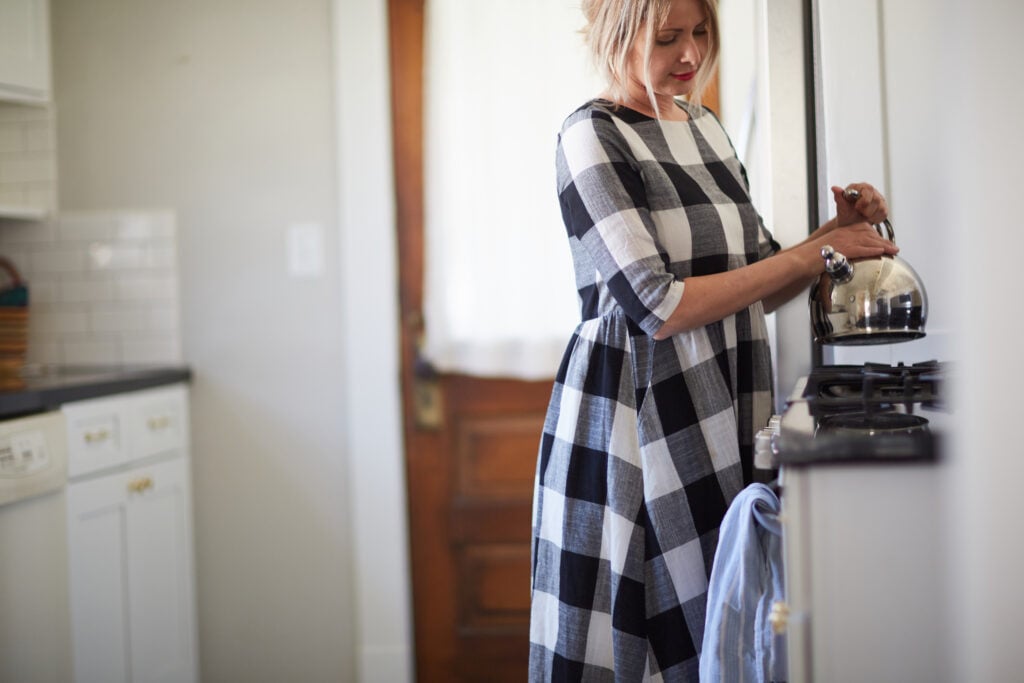 A Christian woman in a navy and white checkered dress setting a stainless steel teapot on the stove as she practices biblical stewardship