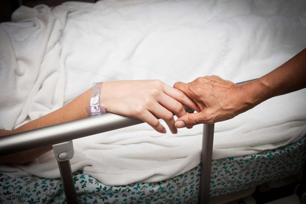 A Christian woman laying in a hospital bed as a loved one reaches to hold her hand