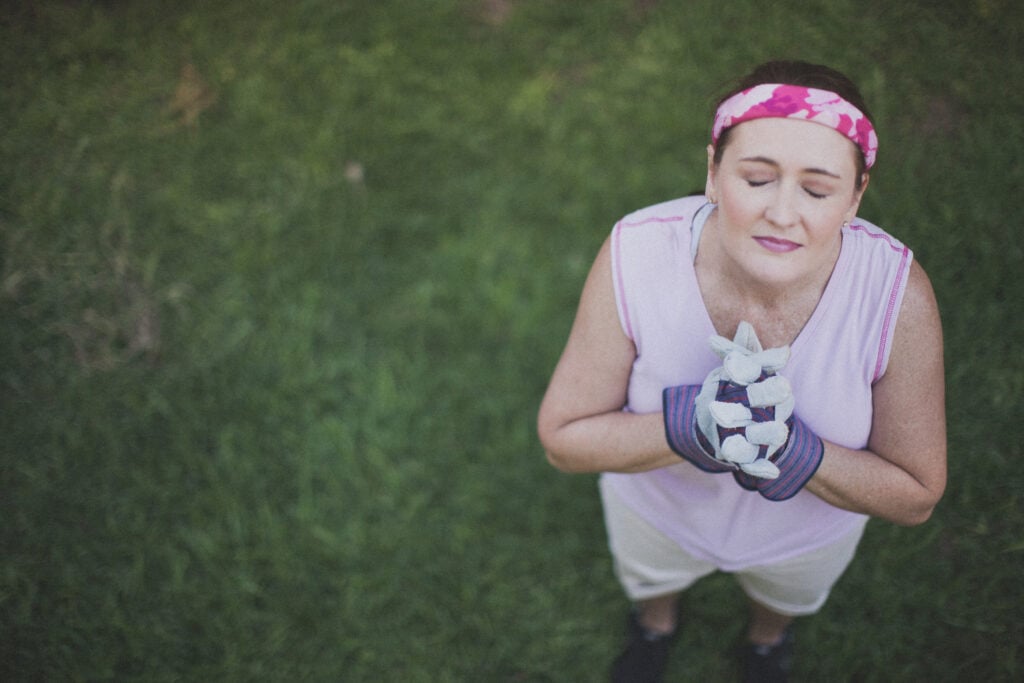 A Christian woman wearing work gloves folding her hands, closing her eyes, and lifting her face in prayer as she practices rhythms of grace