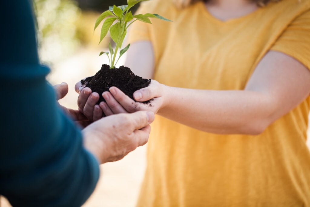 A young Christian woman in a bright yellow t-shirt, cradling a new plant in her hands and extending it to an older person's hands
