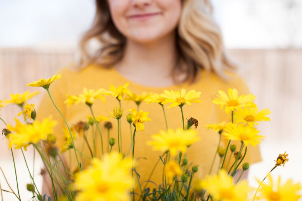 A Christian woman in a bright yellow t-shirt, standing behind a bunch of yellow daisies, and smiling because she is no longer weary of doing good