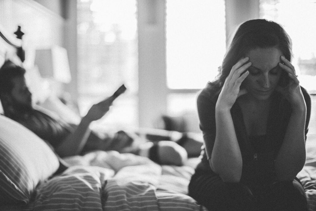 A Christian wife sitting on the edge of the bed with her head in her hands, thinking about how a gentle answer turns away wrath, as her husband lies on the bed with a remote control in his hand
