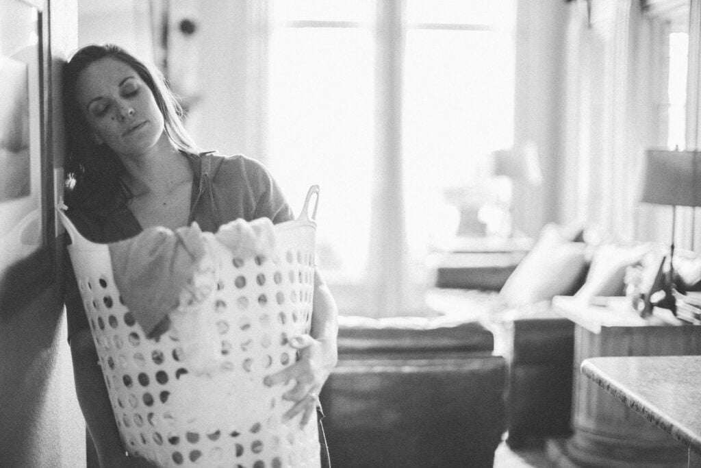 A Christian woman leaning against a wall with her eyes closed while holding a hamper full of laundry, thinking about how a gentle answer turns away wrath