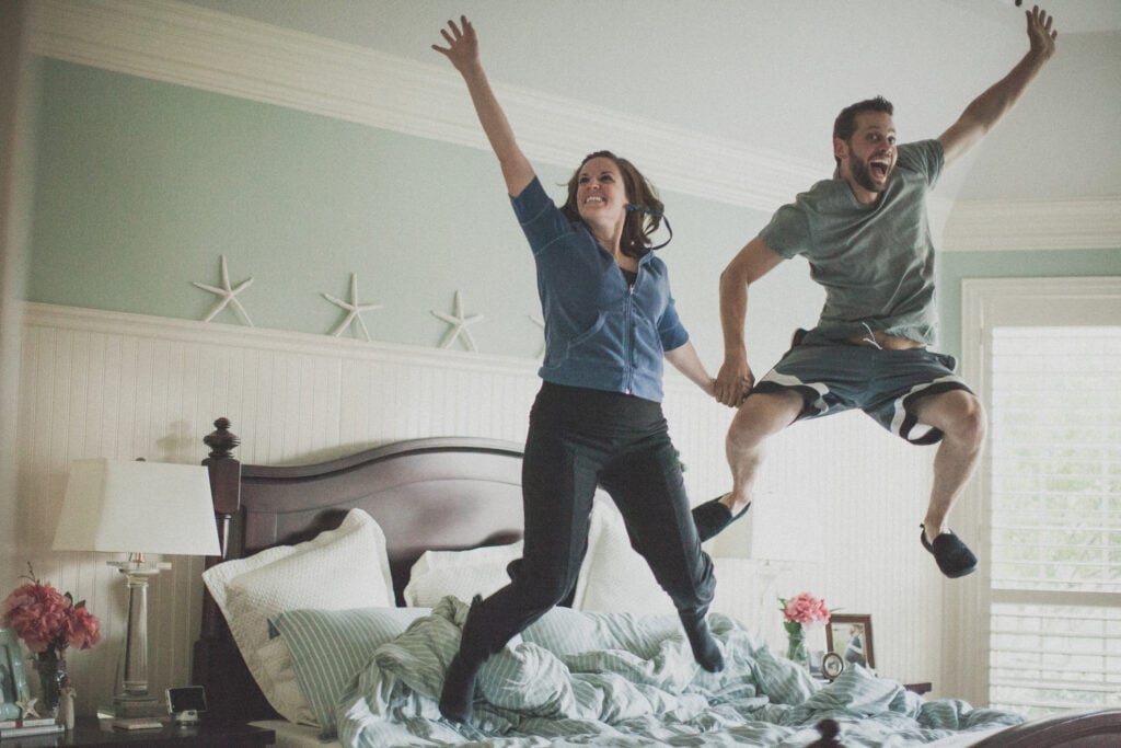 A Christian wife and husband, holding hands and jumping exuberantly on their bed