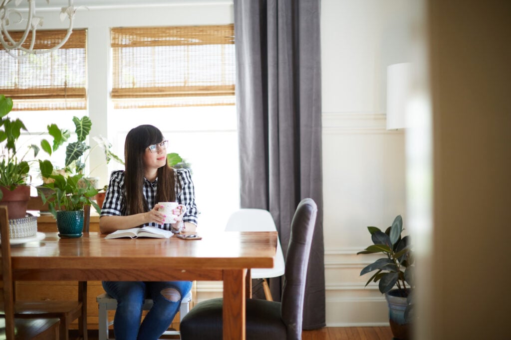 A Christian woman sitting at her dining table with a mug in her hands and her Bible open in front of her, considering the impact of perfectionism in your quiet time